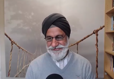 Gurnam Singh on a video call: Man in a light grey v neck jumper with neat, pointy white beard and wearing a dark grey turban against a neutral background and with a bookcase to the right of the screen.